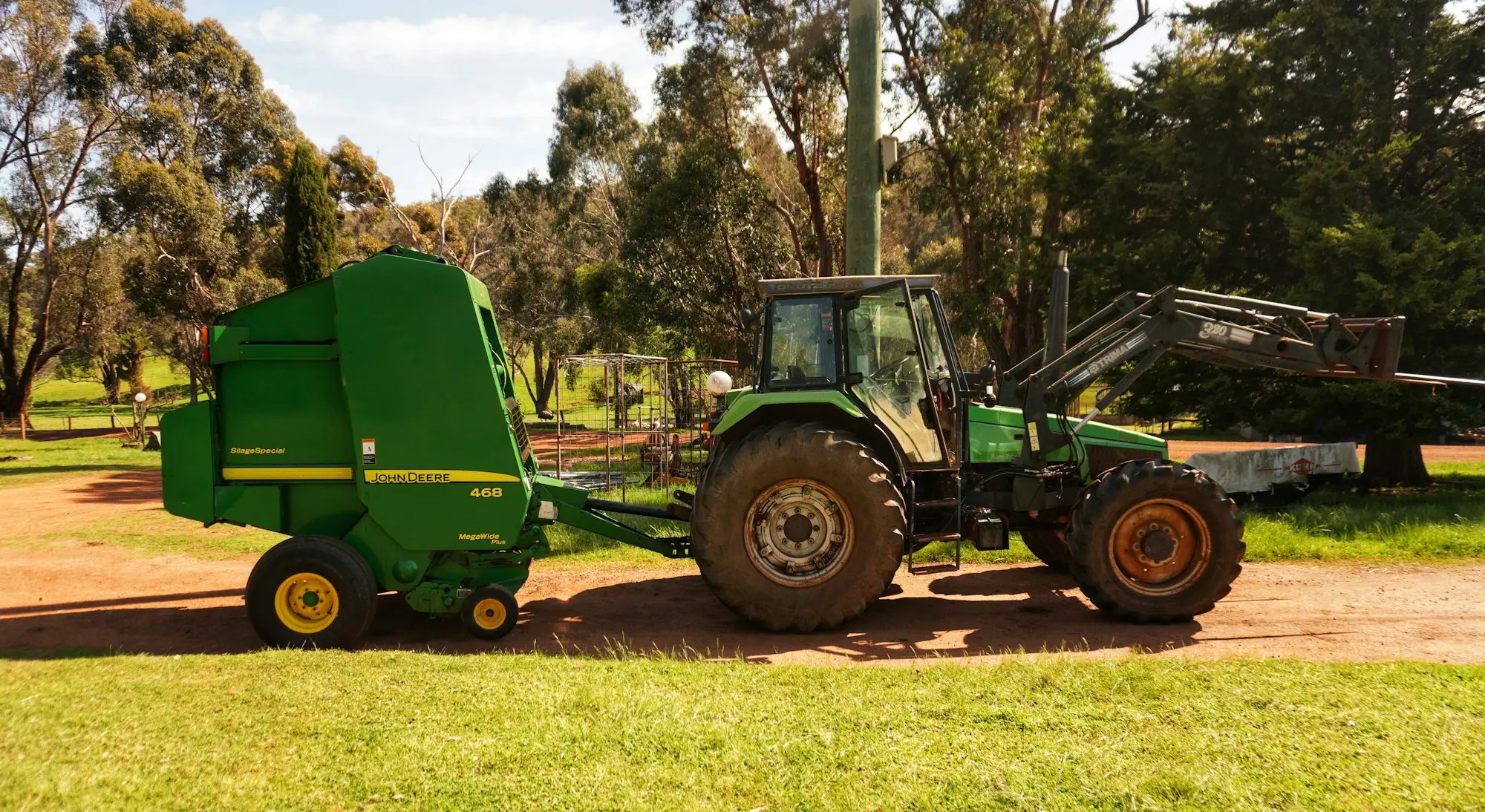 Green tractor towing a baler across a paddock among gum trees
